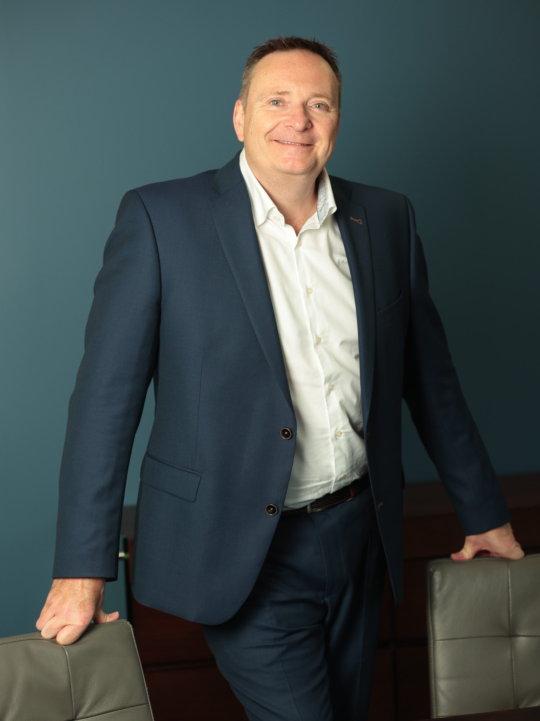 An image of Dante Peters wearing a white shirt and navy suit whilst standing next to a chair.