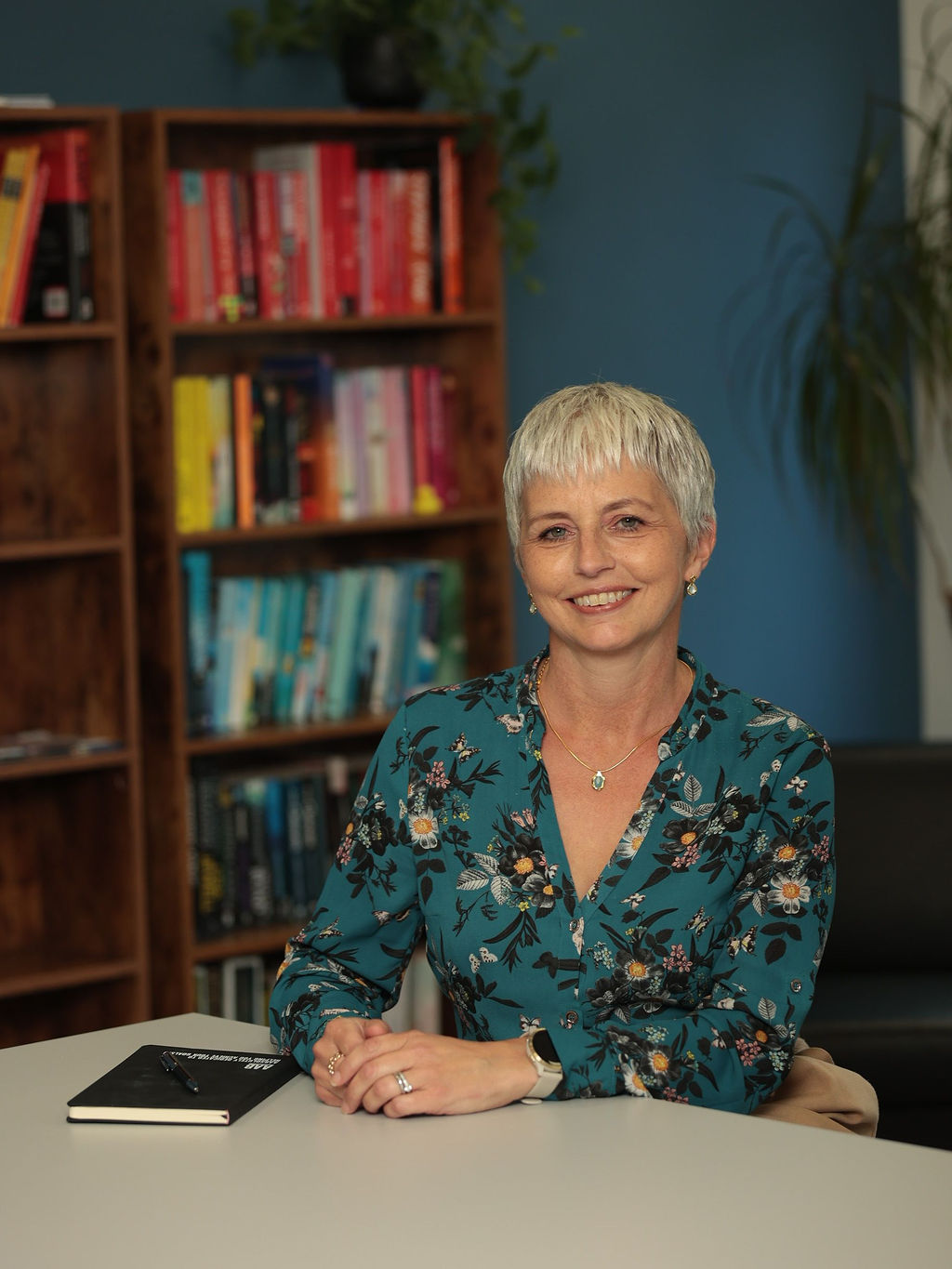 An image of Amanda Mayes sitting at a desk and wearing a blue top with her notebook on the table.