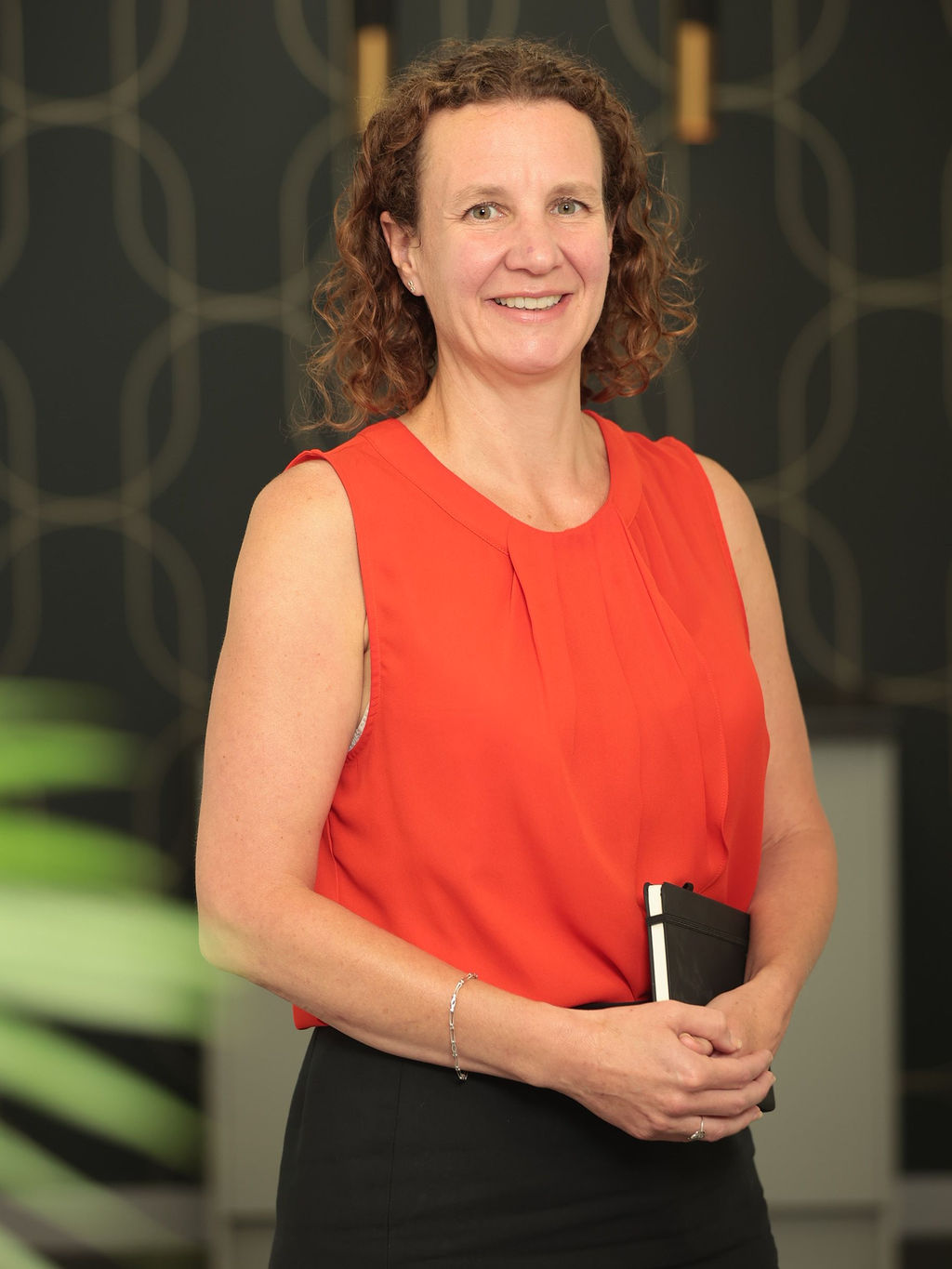 An image of Angela Winton standing in the office and holding a black notebook. She is wearing a red top and black trousers.