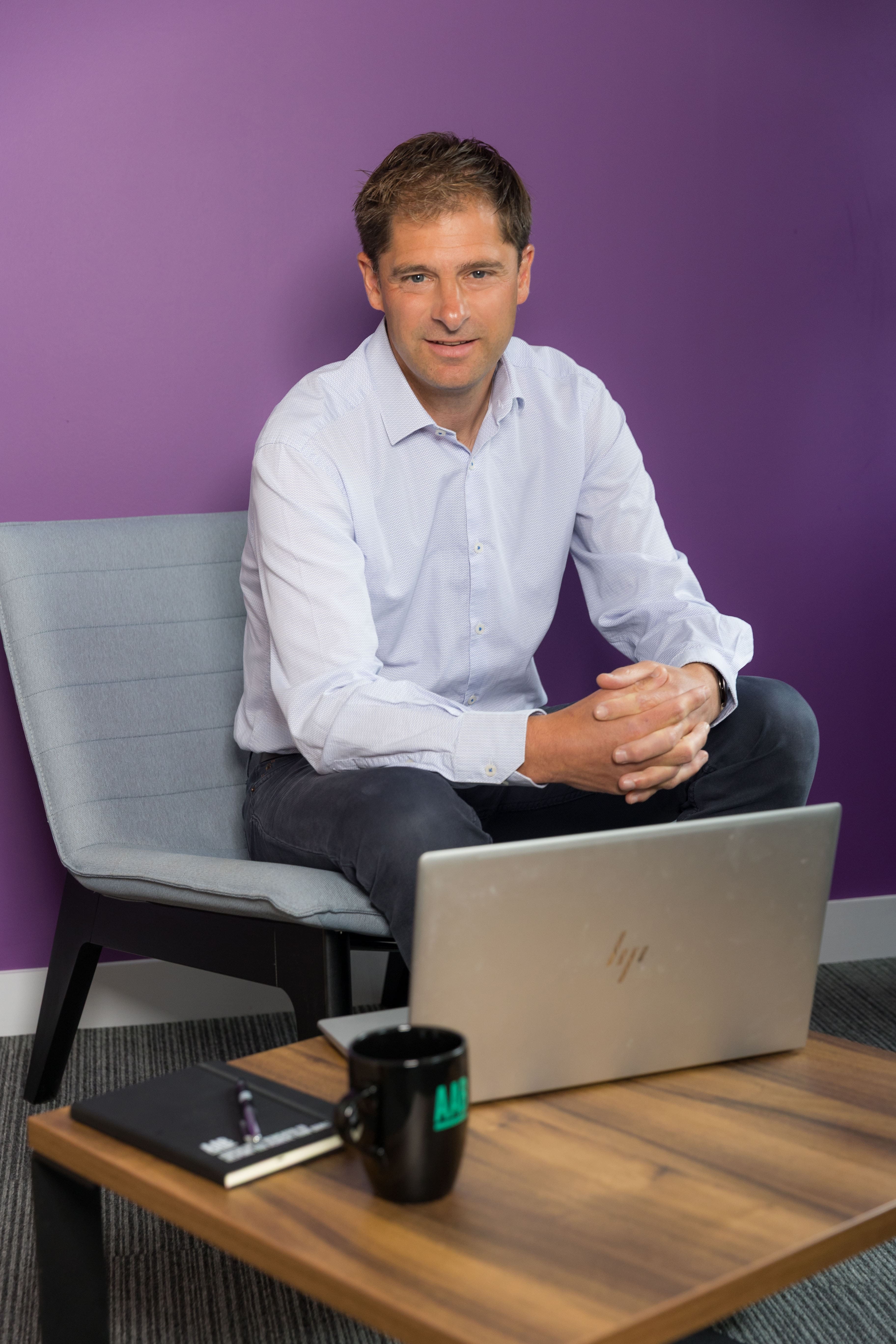An image of Colin Judd sitting on a chair with his laptop and mug on a desk.