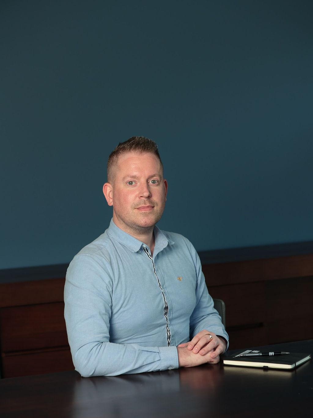 An image of David Felton wearing a blue shirt. He is sitting down and leaning against a table.