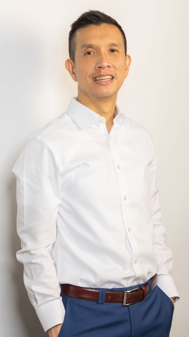 Alex Bui standing smiling at camera with his hands in his pockets against a wall